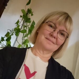 Satu mustassa villatakissa, jonka alla valkoinen Vasemmistoliiton logopaita. Satu hymyilee. Hänen takanaan on kultaköynnös. 